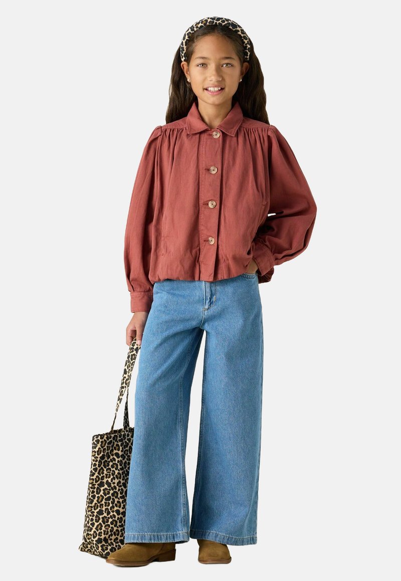 Veste boutonnée couleur rouille avec des manches bouffantes, associée à un jean bleu à jambe large et un sac fourre-tout en léopard. Bottines en suede marron.