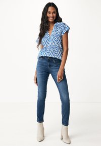 Blusa blu a fantasia con maniche corte, abbinata a jeans blu scuro e stivali con tacco beige. Tessuto morbido con un design a stampa geometrica.