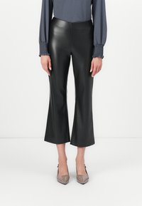 Schwarze, ausgestellte cropped Hosen aus Kunstleder. Der Bund ist glatt und der Stoff hat eine glänzende Oberfläche. Kombiniert mit beigen spitzen Absätzen.