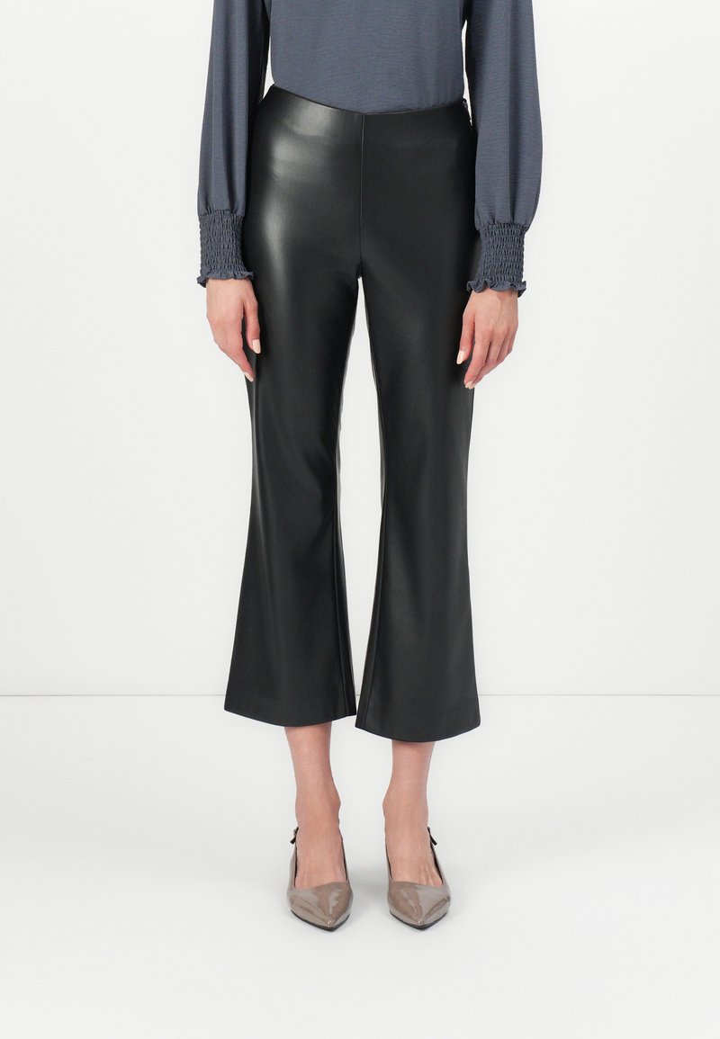 Schwarze, ausgestellte cropped Hosen aus Kunstleder. Der Bund ist glatt und der Stoff hat eine glänzende Oberfläche. Kombiniert mit beigen spitzen Absätzen.
