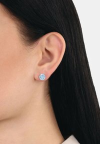 Pendientes de botón redondos con piedras azul claro, montados en metal, que exhiben un diseño facetado. Vista en primer plano junto a la oreja de un modelo.