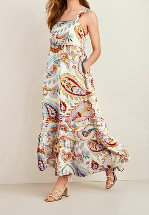 Woman wearing a sleeveless, paisley-patterned maxi dress with side pockets and beige strappy block-heeled sandals on a neutral background.