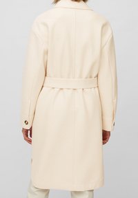 Abrigo beige de mezcla de lana con cintura ceñida, grandes puños y silueta recta, que presenta un acabado limpio y una textura sutil.