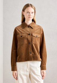 Femme aux cheveux blonds raides, portant une veste en velours côtelé marron avec deux poches sur la poitrine et un pantalon crème à côtes, se tenant devant un fond uni.