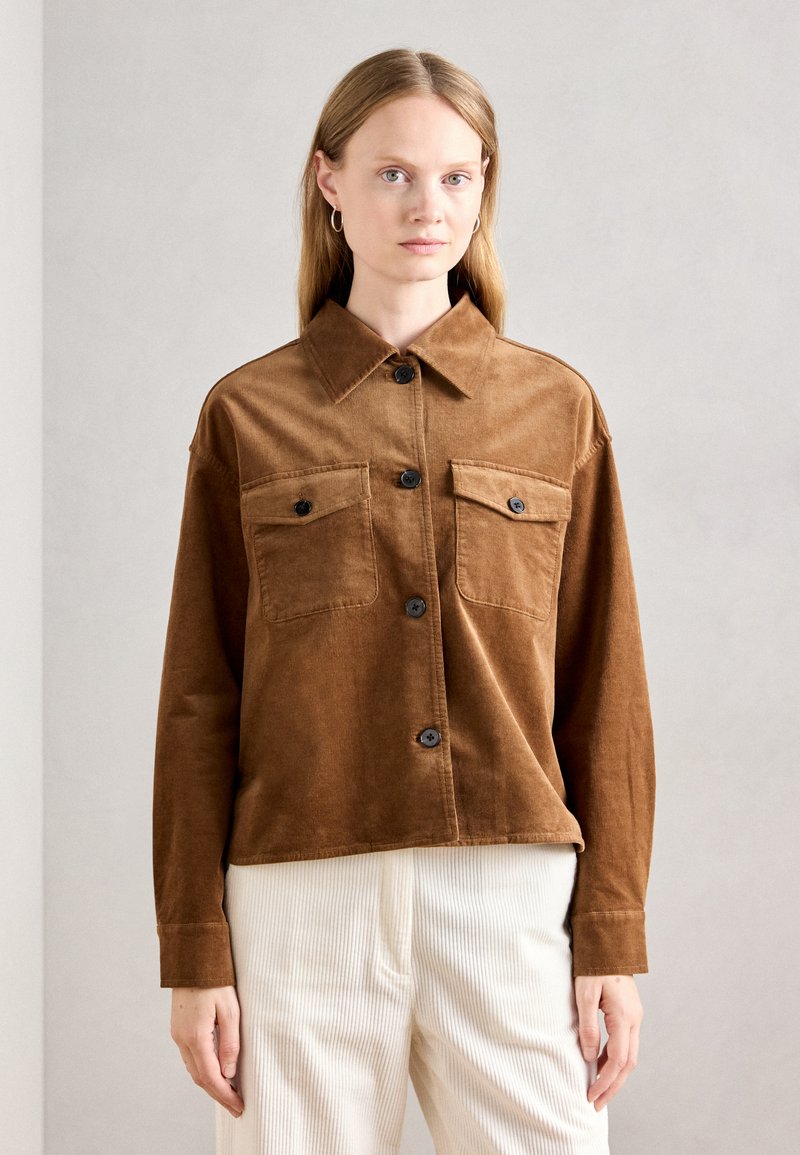 Femme aux cheveux blonds raides, portant une veste en velours côtelé marron avec deux poches sur la poitrine et un pantalon crème à côtes, se tenant devant un fond uni.