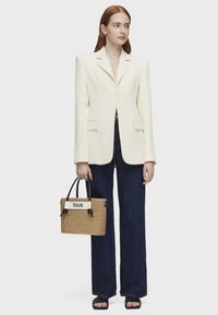 Kremowa marynarka z wyraźnymi ramionami, połączona z ciemnymi spodniami typu wide-leg. Trzyma plecioną, jasnobrązową torbę z czarnymi uchwytami i białym logo.