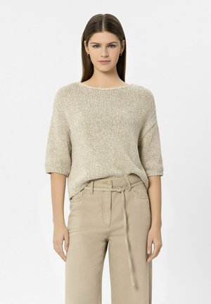 Frau steht und trägt einen beigen kurzärmeligen Strickpullover, der in eine hochtaillierte beige Hose mit gebundenem Gürtel gesteckt ist.