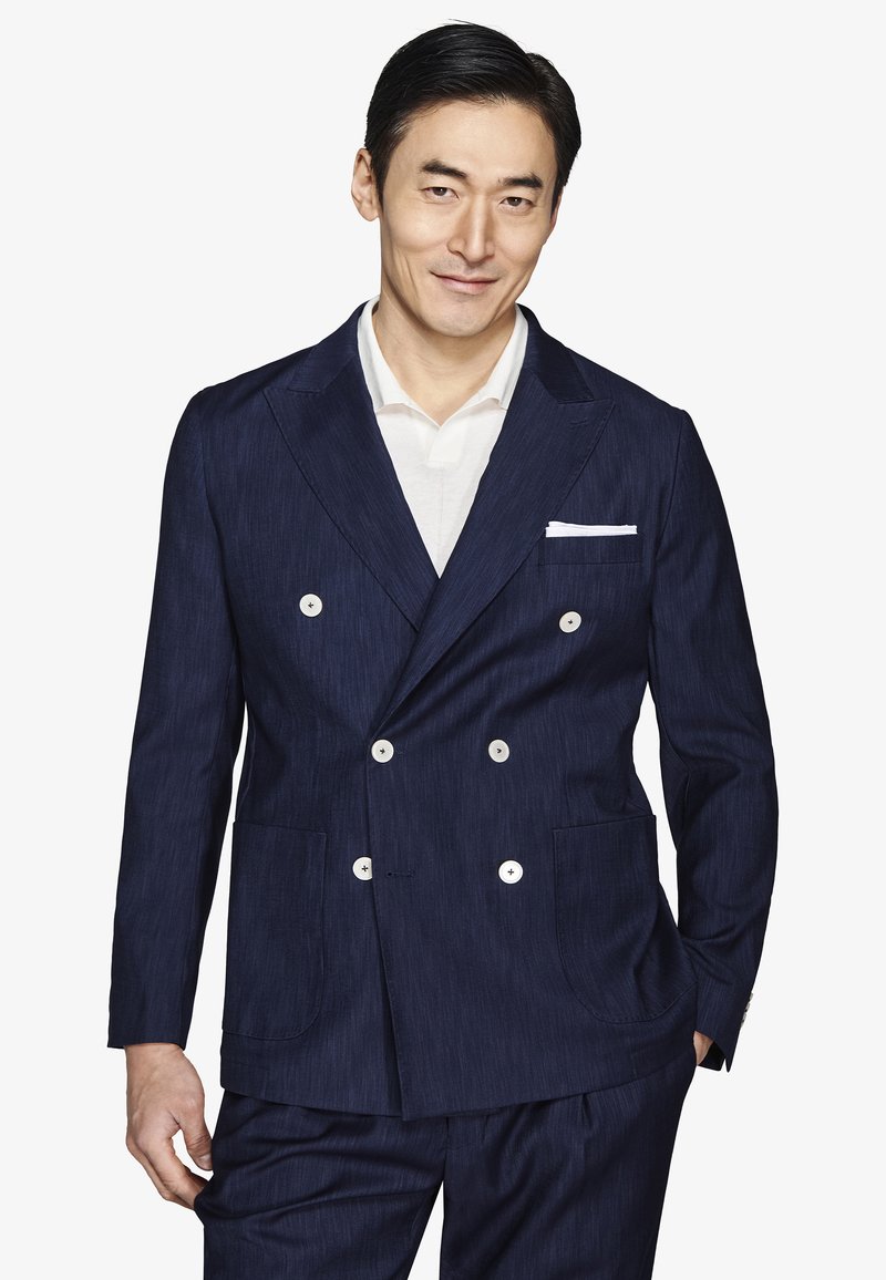 Hombre vestido con un traje azul oscuro de doble botonadura con botones blancos y una camisa blanca, sonriendo, con una mano en el bolsillo.