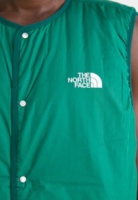 Green sleeveless vest made of lightweight fabric with a smooth texture, featuring snap buttons and a white logo on the left side.