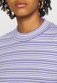 Světle fialový a bílý pruhovaný top s vysokým límcem vyrobený z žebrované látky, s těsným střihem a hladkou texturou. Viditelné zlaté kruhové náušnice.