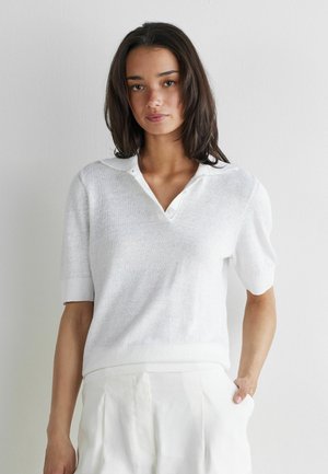 Jeune femme aux cheveux foncés mi-longs portant un pull blanc à manches courtes avec col et un pantalon blanc sur un fond uni.