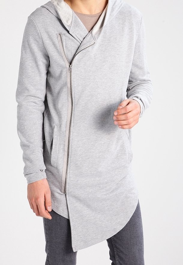 Homme portant un sweat à capuche zippé asymétrique gris clair avec capuche, associé à un pantalon gris foncé, debout devant un fond uni.