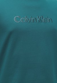 Τυρκουάζ ύφασμα με ανάγλυφο λογότυπο "Calvin Klein" στο κέντρο της θωρακικής περιοχής ενός ενδύματος.