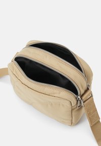 Beżowa torba na ramię z płótna z dwiema zamykanymi na zamek komorami, czarnym wnętrzem i regulowanym paskiem. Posiada srebrne okucia i subtelne detale przeszycia.