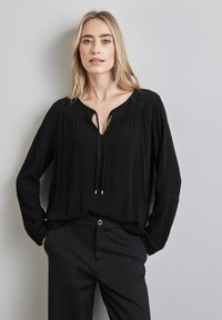 Zwarte blouse met lange mouwen, een V-hals en een koorddetail, met subtiele plooien en een gestructureerde stof. Geselecteerd met zwarte broeken.
