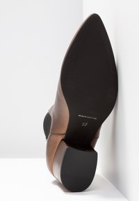 Chaussure en cuir marron à bout pointu avec semelle noire et talon bloc, taille 37, marquée "Made in Portugal,", photographiée par en dessous contre un mur en angle.