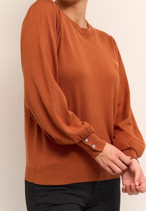 Jersey de manga larga color naranja óxido con mangas abullonadas y dos botones decorativos de cristal en el puño, combinado con pantalones negros.