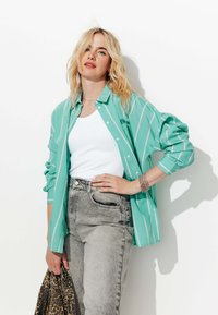 Chemise à rayures turquoise sur un débardeur blanc, associée à un jean taille haute gris. Accessoirisée avec un sac à motifs et des bracelets.