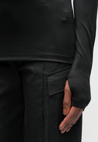 Bluza neagră cu mânecă lungă, realizată dintr-un material neted, având o deschidere pentru degetul mare la manșetă, purtată împreună cu o pereche de pantaloni negri care au o buzunar lateral.
