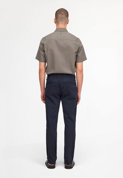 Homme debout, dos tourné, portant une chemise grise à manches courtes, un pantalon bleu marine et des chaussures noires sur un fond blanc uni.