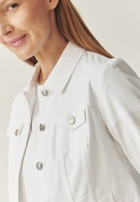 Veste en denim blanche avec un col, deux poches poitrine et des éléments en métal argenté. Présente une texture côtelée et des fermetures à bouton.