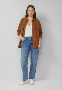 Bruine suède blouse met knopen over een wit tanktopje, gecombineerd met lichtblauwe spijkerbroek en witte sneakers. Heeft twee borstzakken.