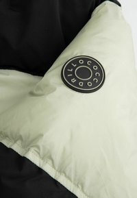 Kurtka typu puffer z czarną górną częścią i jasnoskalnym dolnym fragmentem. Posiada okrągłą czarną naszywkę z logo na dolnej części ramienia.