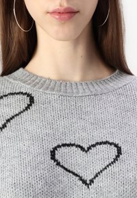 Detailní záběr dolní části obli�čeje a krku ženy, která má na sobě stříbrné kruhové náušnice a šedý svetr s černými srdíčky.