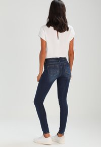 Jeans skinny de mezclilla oscura con cintura alta, que cuentan con bolsillos traseros y costuras sutiles. Combinados con una blusa blanca de manga corta.