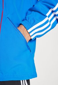 Modra športna jakna z elastičnimi manšetami, belimi črtami na rokavih in stranskim žepom. Gladka tekstura tkanine. Detajl z zadrgo na žepu.