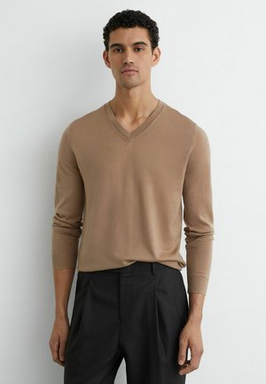 Beżowy sweter z dekoltem w serek z miękkiej dzianiny z długimi rękawami, w połączeniu z ciemnymi spodniami. Prosty fason, gładka faktura, minimalne zdobienia.