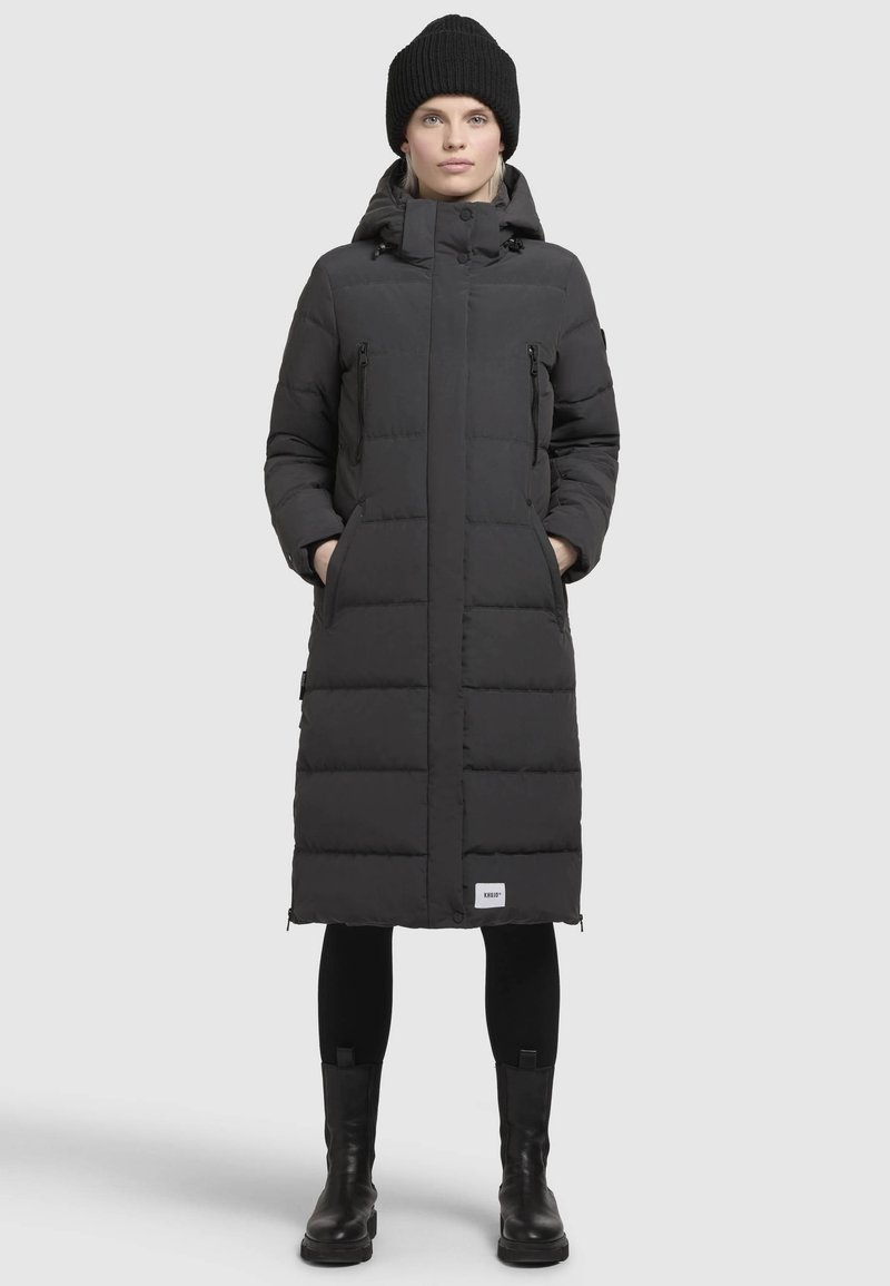 Black puffer coat with a knee-length design, zippered pockets, high collar, and hood. Features horizontal quilted pattern. Worn with black leggings and boots.
