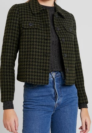Kobieta ubrana w zielono-czarną kraciastą, przyciętą kurtkę na guziki, na ciemnoszarym topie i niebieskie dżinsy z wysokim stanem.