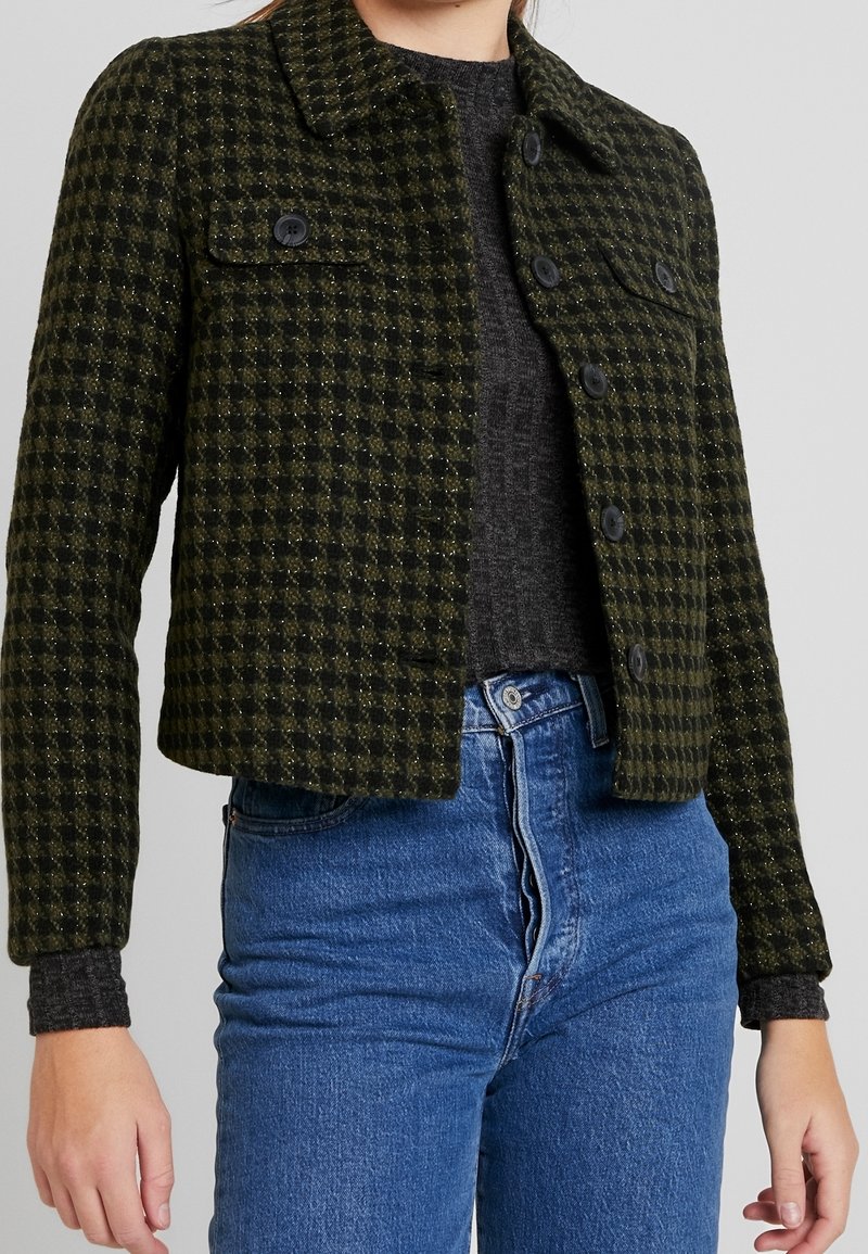 Femme portant une veste courte à carreaux verts et noirs boutonnée par-dessus un haut gris foncé et un jean taille haute bleu.