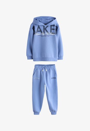 Teenager iført blå "Baker by Ted Baker" hoodie og matchende joggingbukser, står med den ene hånd i lommen og hvide sneakers på gråt gulv.