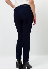 Jeans skinny azul oscuro con textura suave, con bolsillos traseros y una cintura alta. Llevados con botas negras hasta el tobillo.