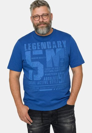 Bärtiger Mann mit Brille, der ein blaues T-Shirt mit dem Schriftzug „Legendary 57“ und schwarze abgenutzte Jeans trägt, steht mit den Händen in den Taschen.