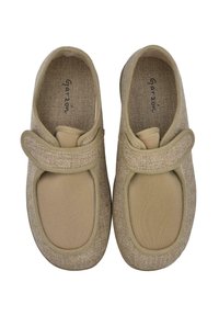 Zapatillas beige de tipo slip-on con parte superior de tela suave, punta redonda y correa de velcro. Presentan detalles texturizados y una plantilla acolchada.
