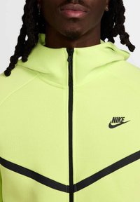 Chaqueta Nike en amarillo neón, con cierre frontal, acentos en negro y rayas contrastantes en negro a lo largo del pecho. Textura suave y diseño ajustado.