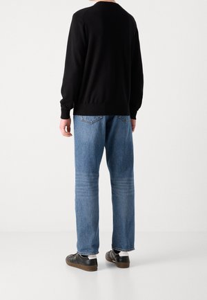 Person trägt ein schwarzes Langarmoberteil, blaue Jeans und schwarze Turnschuhe, steht mit dem Rücken zur Kamera auf einem einfarbigen hellen Hintergrund.