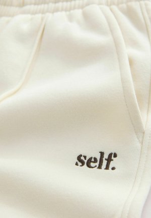 Bēšas sporta bikses ar brīvu piegriezumu, elastīgu jostu, kabatām un mazu melnu "self." logotipu kreisajā pusē. Mīksta tekstūra.