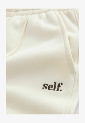 Beiget collegehousut, joissa on rento istuvuus, joustava vyötärönauha, taskut ja pieni musta "self."-logo vasemmalla puolella. Pehmeä pinta.