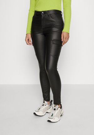 Pantalones negros de imitación cuero con corte slim, que cuentan con bolsillos cargo y cremalleras en los tobillos, combinados con zapatillas deportivas grises y blancas.