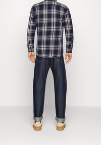 Rutig skjorta i marinblått och krämvitt, långärmad med rundad nederkant. Mörkblå jeans med uppvikta fållar, kombinerat med beige skor.