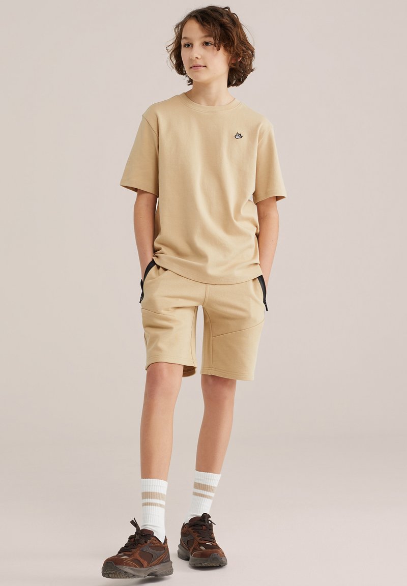 Enfant aux cheveux bouclés mi-longs portant un t-shirt beige et un short, des chaussettes blanches et des baskets marron, debout les mains dans les poches.