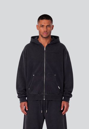 Man staat, draagt een zwarte hoodie met ritssluiting en voorzakken en bijpassende zwarte broek tegen een effen grijze achtergrond.