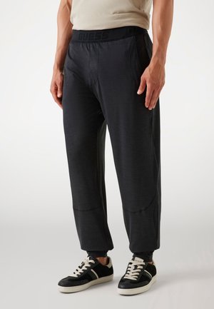 Man draagt zwarte Guess joggingbroek en zwart-witte sneakers, staand tegen een effen achtergrond.