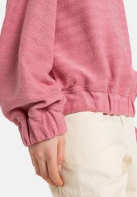 Pink textured sweatshirt featuring a herringbone pattern, elastic cuffs, and a gathered hem. Worn over light-colored pants.
