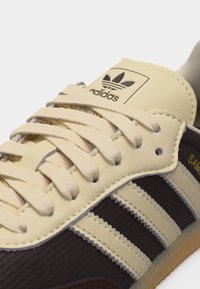 Beige ja tumepruuni Adidas tennise lähipilt, paeltega, kolme Adidas triibu ja küljele trükitud „Samba“ tekstiga.