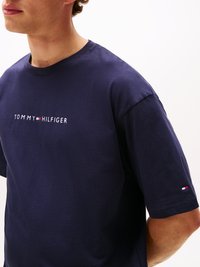 Tumši zils kokvilnas T-krekls ar īsām piedurknēm, kurā ir balts "TOMMY HILFIGER" logo un sarkans un balts akcents uz piedurknes.